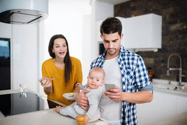 A portrait of young family standing in a kitchen at home, a man with smartphone holding a baby and a woman feeding her with a spoon. - HPIF31038