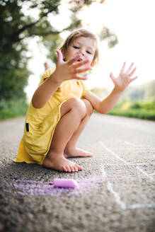 A small cute girl on a road in countryside in sunny summer nature, drawing with chalk. - HPIF31039