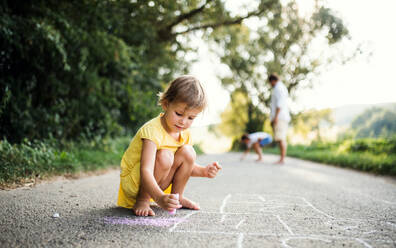 A small cute girl on a road in countryside in sunny summer nature, drawing with chalk. - HPIF31041