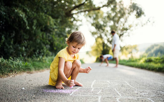 A small cute girl on a road in countryside in sunny summer nature, drawing with chalk. - HPIF31041