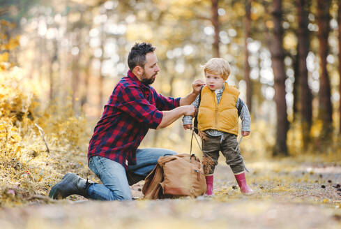 A mature father putting a backpack on a toddler son on a road in an autumn forest. - HPIF31114