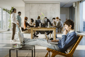 Side view of young businessman using laptop in chair while colleagues in background at creative office - MASF36983