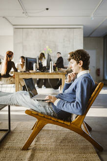 Young businessman using laptop on chair while colleagues in background at creative office - MASF36984