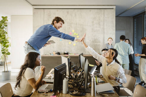 Cheerful entrepreneurs giving high-five while colleagues sitting at desk in creative office - MASF36992