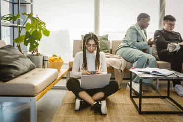 Programmer using mobile phone while sitting with laptop on carpet in creative office - MASF37000