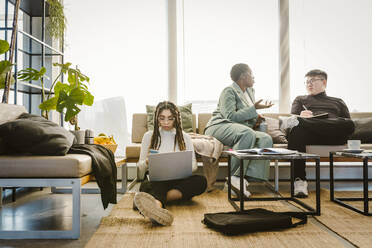 Programmer using mobile phone sitting on carpet while colleague discussing in creative office - MASF37002