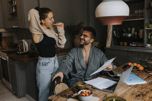 Non-binary person brushing teeth while talking to man sitting at dining table in home - MASF37020