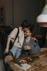 Non-binary person kissing boyfriend sitting at dining table in room - MASF37022