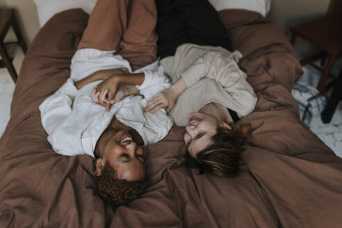 Smiling non-binary couple holding hands while lying down on bed at home - MASF37045