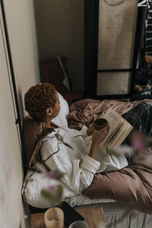 High angle view of non-binary person reading book while sitting on bed at home - MASF37054