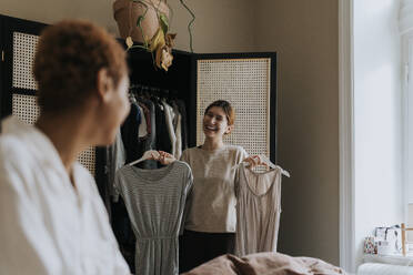 Happy non-binary person holding dress while standing near wardrobe at home - MASF37058