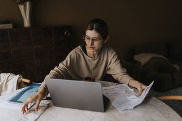 Non-binary person collecting bills while sitting with laptop at home - MASF37072