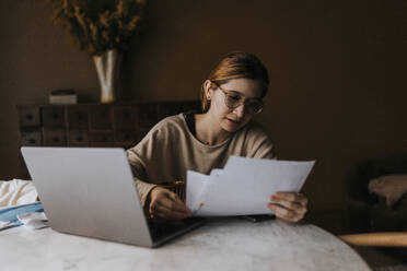 Non-binary person reviewing bills by laptop on table at home - MASF37073