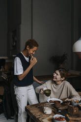 Smiling non-binary person brushing teeth by friend having breakfast at home - MASF37088