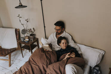 High angle view of man reading book by non-binary person using mobile phone on bed at home - MASF37098
