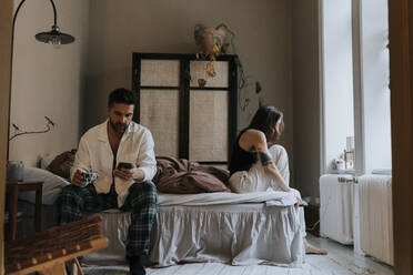 Man using smart phone with non-binary person wearing socks sitting on bed at home - MASF37103