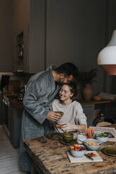 Man kissing smiling non-binary person while having breakfast at home - MASF37113