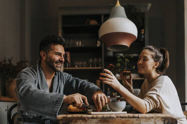 Happy non-binary person sharing smart phone with boyfriend while having breakfast at home - MASF37116