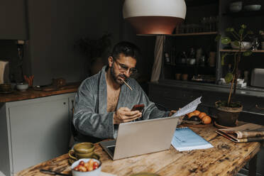 Man holding bills using smart phone while sitting with laptop on dining table at home - MASF37123