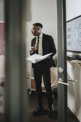 Mature male entrepreneur holding notebook during business meeting at office seen through doorway - MASF37165