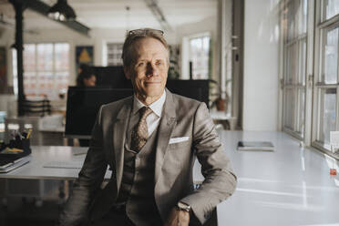 Smiling mature businessman wearing suit sitting by desk at office - MASF37171