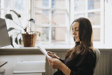 Side view of businesswoman examining document sitting at office - MASF37172