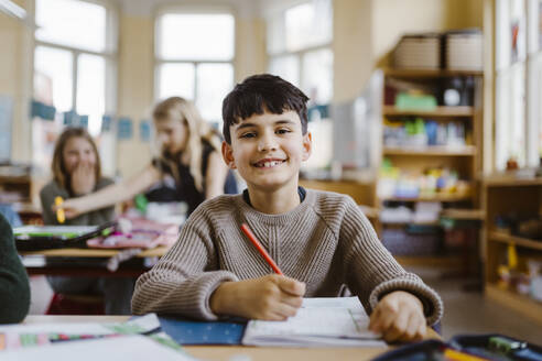 Portrait of happy male pupil sitting at desk in classroom - MASF37220