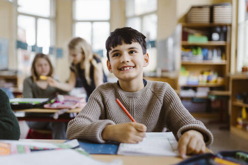 Smiling male pupil holding pencil while sitting at desk in classroom - MASF37221
