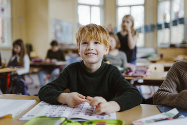 Smiling male pupil with blond hair sitting at desk in classroom - MASF37222