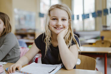 Smiling blond schoolgirl leaning on elbow while sitting at desk in classroom - MASF37223