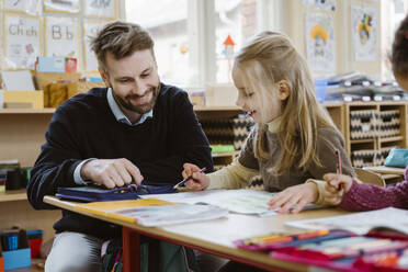 Smiling male teacher assisting female student while studying in classroom - MASF37230