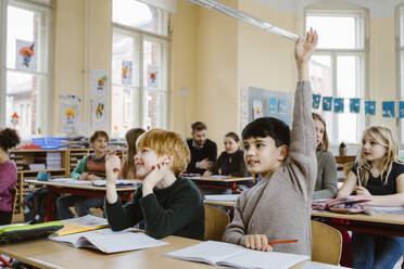 Boy with hand raised answering during lecture while sitting by male friend in classroom - MASF37236