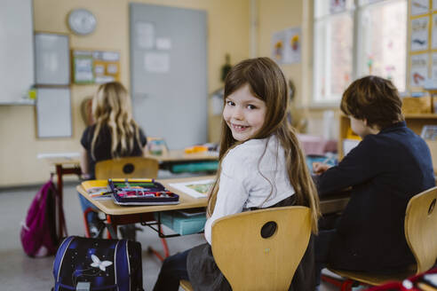 Portrait of smiling girl looking over shoulder sitting on chair by friend in classroom - MASF37237