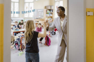 Smiling female teacher and schoolgirl giving high-five at doorway in classroom - MASF37246