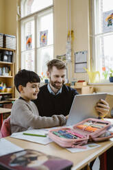 Happy male teacher assisting schoolboy using tablet PC at desk in school - MASF37247
