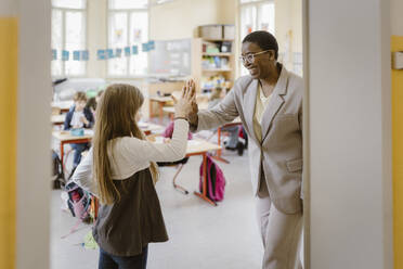 Happy teacher giving high-five to schoolgirl standing at doorway in school - MASF37250