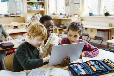 Happy teacher assisting boy and girl with digital tablet sitting at desk in classroom - MASF37252