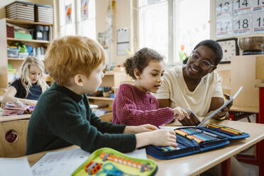 Smiling teacher assisting boy and girl with digital tablet sitting at desk in classroom - MASF37254