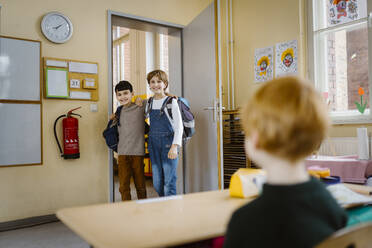 Happy schoolboys with arms around entering classroom and looking at friend - MASF37255