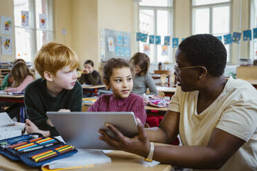 Female teacher assisting schoolboy and schoolgirl with digital tablet in classroom - MASF37256