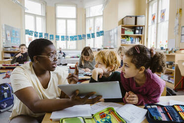 Teacher with tablet PC explaining schoolboy and schoolgirl at desk while gesturing in classroom - MASF37258