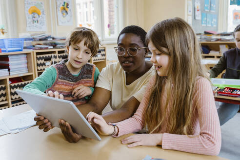 Female teacher sitting amidst schoolboy and schoolgirl using digital tablet sitting at desk in classroom - MASF37261