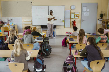 Happy female teacher standing against whiteboard with students in classroom - MASF37263