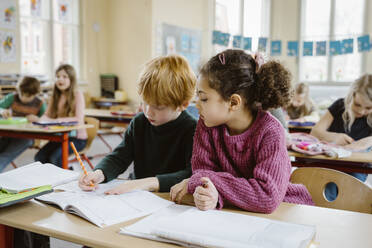 Girl sitting besides blond boy writing in book at desk in classroom - MASF37264