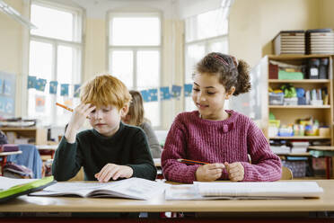 Smiling girl sitting by confused blond boy at desk in classroom - MASF37266