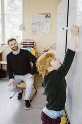 Blond boy writing on whiteboard near male teacher sitting in classroom - MASF37267