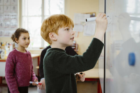 Side view of blond boy writing on whiteboard while solving mathematics in classroom - MASF37268