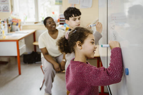 Girl and boy writing on whiteboard while solving mathematics in classroom - MASF37269