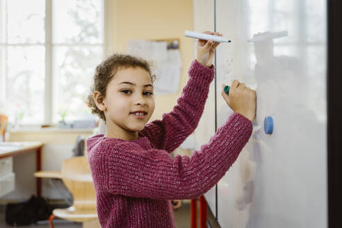 Portrait of smiling girl writing on whiteboard in classroom - MASF37271