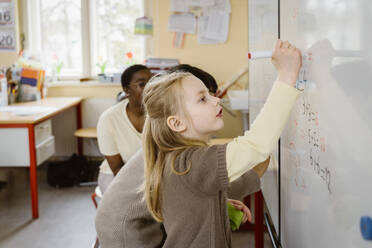 Blond girl solving maths problem while writing on whiteboard in classroom - MASF37272